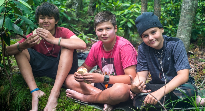 Three people sit in a wooded area holding sandwiches 